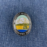 An oval enamel Walking Stick Medallion featuring a mountain landscape with the text 'LASSEN VOLCANIC NATIONAL PARK' on a blue denim background.