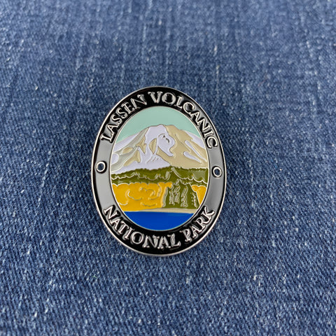 An oval enamel Walking Stick Medallion featuring a mountain landscape with the text 'LASSEN VOLCANIC NATIONAL PARK' on a blue denim background.