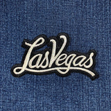 An embroidered script patch that reads "Las Vegas" in shimmering silver  thread with a black outline on denim.