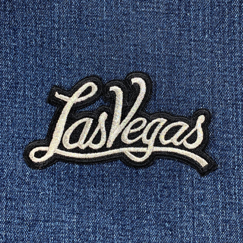 An embroidered script patch that reads "Las Vegas" in shimmering silver  thread with a black outline on denim.