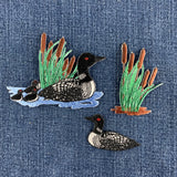 Three embroidered loon and cattail patches, displayed on a blue denim background.