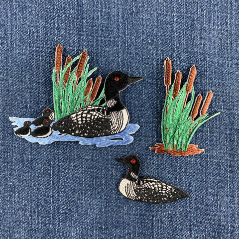Three embroidered loon and cattail patches, displayed on a blue denim background.