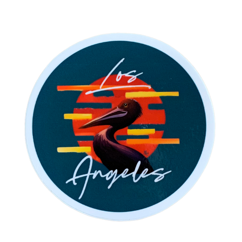 Circular sticker with a dark teal background. The design features a stylized brown pelican silhouette centered over an orange-red circle resembling a sunset, with horizontal yellow and orange stripes. White cursive text above and below the pelican reads "Los Angeles."