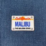 Embroidered patch shaped like a California license plate, featuring the word 'MALIBU' in large blue letters, 'California' and a setting sun in orange and red above, and 'THE GOLDEN STATE' in black below, all on a white background on blue denim.