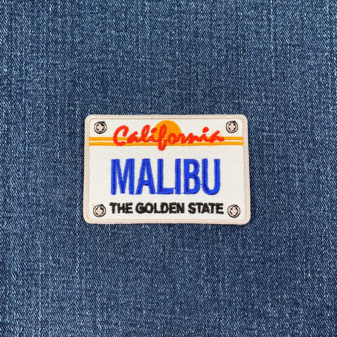 Embroidered patch shaped like a California license plate, featuring the word 'MALIBU' in large blue letters, 'California' and a setting sun in orange and red above, and 'THE GOLDEN STATE' in black below, all on a white background on blue denim.
