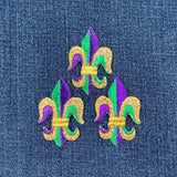 Three embroidered fleur-de-lis applique patches in purple and green with metallic gold accents, designed for iron-on application on a denim background