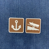 Two square embroidered patches with rounded corners on a blue denim fabric background. The left patch is brown with a white anchor symbol, and the right patch is brown with a white boat and trailer symbol.