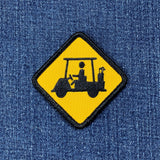 A yellow diamond-shaped embroidered patch with a black border and a black silhouette of a golf cart driver, shown on a blue denim fabric background.