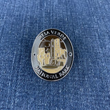 A round enamel Walking Stick Medallion featuring a silver-toned border with the text 'MESA VERDE NATIONAL PARK' and a gold-toned central illustration of mesa formations, shown on a blue denim background.
