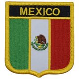 An embroidered shield-shaped patch featuring the Mexican flag with green, white, and red vertical stripes, the Mexican coat of arms in the center, and the word 'MEXICO' in yellow embroidery on a black banner at the top, all outlined with a gold border.