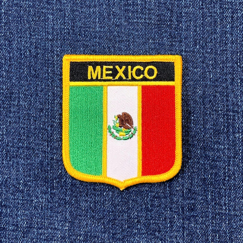An embroidered shield-shaped patch featuring the Mexican flag with 'MEXICO' in yellow text on a black background, placed on a blue denim fabric surface.