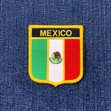 An embroidered shield-shaped patch featuring the Mexican flag with 'MEXICO' in yellow text on a black background, placed on a blue denim fabric surface.