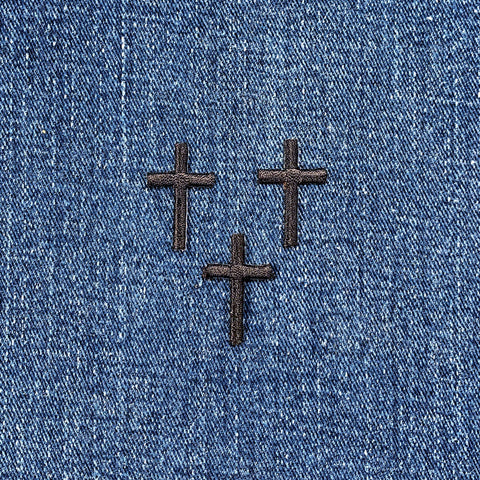 Three black cross applique patches with an embroidered design, intended for iron-on application, on a denim background