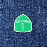 A green and white embroidered patch shaped like a highway sign, reading 'CALIFORNIA 1', placed on a blue denim fabric background.