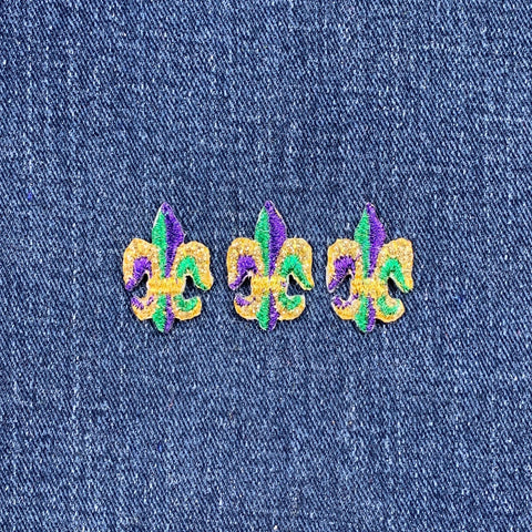 Three Fleur de Lis applique patches in green, gold, and purple Mardi Gras colors on a denim background