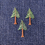 Three embroidered applique patches depicting evergreen trees, with brown trunks and green foliage, displayed on a denim background