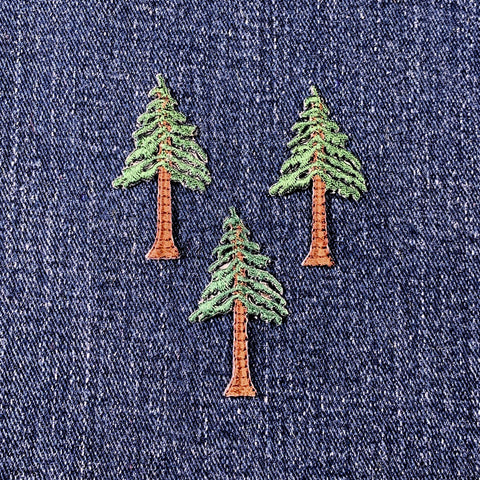 Three embroidered applique patches depicting evergreen trees, with brown trunks and green foliage, displayed on a denim background