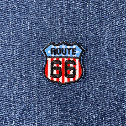 A shield-shaped embroidered patch with the text 'ROUTE 66' on a background resembling the United States of America flag, displayed on a denim background