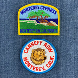 Two embroidered patches displayed on a blue denim background. The top patch features a green cypress tree, brown trunk, blue sky, and the text 'MONTEREY CYPRESS' and 'MONTEREY BAY, CALIF.' and the bottom patch features a sea otter and 'Cannery Row' with 'Monterey, Calif.' below.