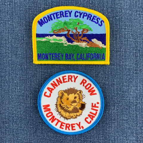 Two embroidered patches displayed on a blue denim background. The top patch features a green cypress tree, brown trunk, blue sky, and the text 'MONTEREY CYPRESS' and 'MONTEREY BAY, CALIF.' and the bottom patch features a sea otter and 'Cannery Row' with 'Monterey, Calif.' below.