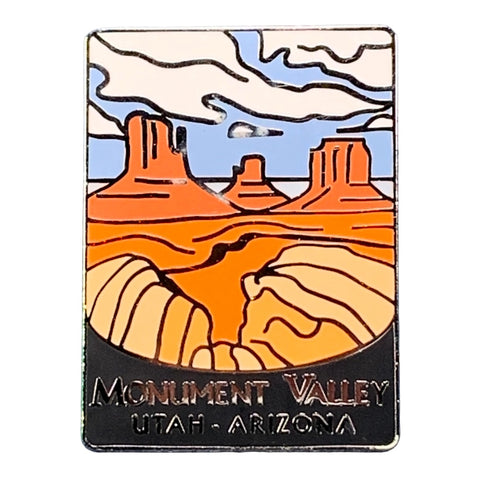 A rectangular enamel pin featuring a stylized Monument Valley landscape with orange rock formations, beige dunes, blue sky, and white clouds. The bottom reads 'MONUMENT VALLEY UTAH - ARIZONA' on a black background.