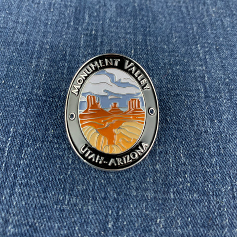 An oval enamel Walking Stick Medallion featuring a scenic Monument Valley landscape with orange rock formations, blue sky, and white clouds, bordered by silver metal with the text 'MONUMENT VALLEY' and 'UTAH - ARIZONA'. The pin is placed on a blue denim fabric background.