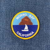 Round patch of a sailboat on dark blue water with Morro Rock, a large brown dome-shaped hill, in the background. White text reads: "MORRO BAY CALIFORNIA."