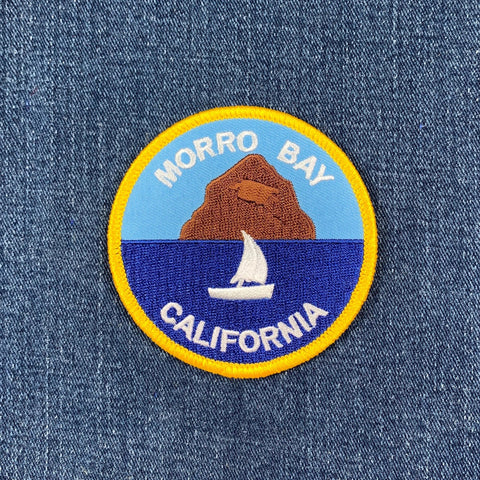 Round patch of a sailboat on dark blue water with Morro Rock, a large brown dome-shaped hill, in the background. White text reads: "MORRO BAY CALIFORNIA."