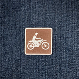 A square brown patch with a white embroidered motorcycle rider icon, shown on a dark blue denim fabric background.