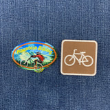 Two bicycle-themed patches displayed on a blue denim background: an oval multicolor patch with a mountain biker and the words 'Mountain Biking,' and a square tan patch with a white bicycle symbol.