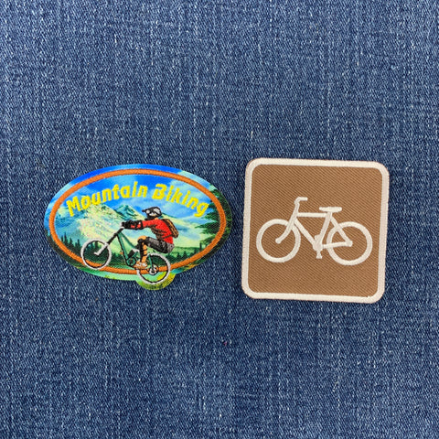 Two bicycle-themed patches displayed on a blue denim background: an oval multicolor patch with a mountain biker and the words 'Mountain Biking,' and a square tan patch with a white bicycle symbol.