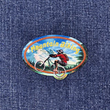 An oval embroidered patch featuring a mountain biker and the words 'Mountain Biking' in yellow, placed on a blue denim fabric background.