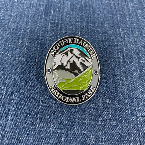An oval metal enamel Walking Stick Medallion pin featuring a mountain landscape with the text 'MOUNT RAINIER NATIONAL PARK,' placed on blue denim fabric.