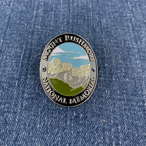 An oval enamel Walking Stick Medallion featuring Mount Rushmore National Memorial, displayed on a blue denim fabric background.