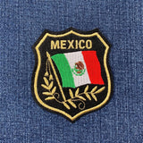 An embroidered shield-shaped patch featuring the word 'MEXICO' above a Mexican flag with a gold laurel design, placed on a blue denim fabric background.