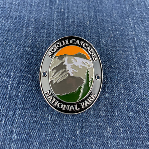 An oval enamel Walking Stick Medallion featuring a mountain landscape with snow, green trees, and an orange sky, bordered by the text 'NORTH CASCADES NATIONAL PARK' on a blue denim background.