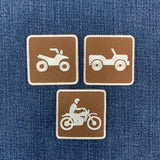 Three square brown embroidered patches with white outlines and vehicle icons, shown on a blue denim background. The top-left patch shows an ATV, the top-right shows an off-road vehicle, and the bottom patch shows a motorcycle with a rider.