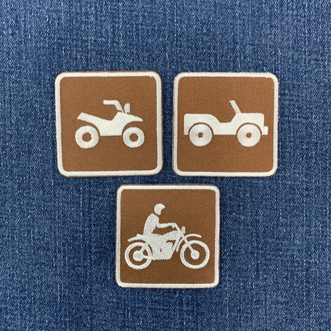 Three square brown embroidered patches with white outlines and vehicle icons, shown on a blue denim background. The top-left patch shows an ATV, the top-right shows an off-road vehicle, and the bottom patch shows a motorcycle with a rider.