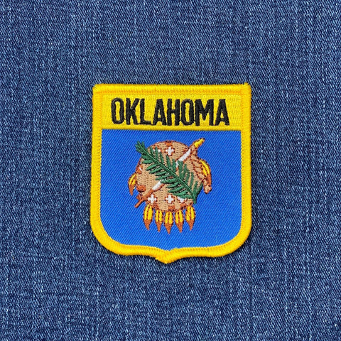 A shield-shaped, embroidered patch with a yellow border and the word "OKLAHOMA" stitched in black on a yellow background at the top on a denim background.