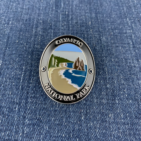 An oval enamel Walking Stick Medallion featuring a landscape of Olympic National Park, with blue water, green hills, and a sandy beach, bordered by a silver-toned metal frame with the text 'OLYMPIC NATIONAL PARK'. The pin is placed on a blue denim background.