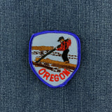 A shield-shaped embroidered patch with a blue border and a depiction of a lumberjack standing on a log with the word 'OREGON' below and displayed on a denim background