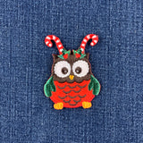 A festive embroidered owl patch with candy cane antennae and a holly crown, shown on a blue denim fabric background.