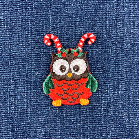 A festive embroidered owl patch with candy cane antennae and a holly crown, shown on a blue denim fabric background.