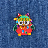 An embroidered owl patch with colorful Christmas lights draped around it, placed on a blue denim fabric background.