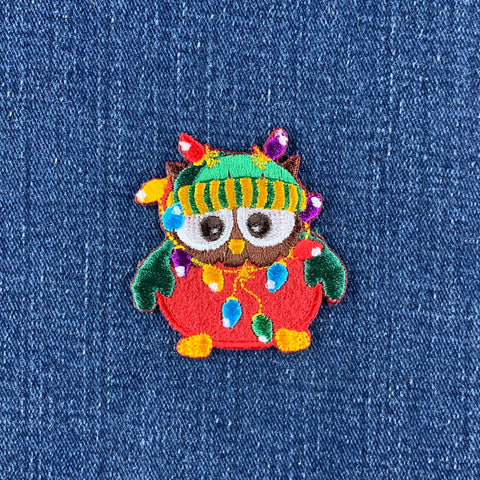 An embroidered owl patch with colorful Christmas lights draped around it, placed on a blue denim fabric background.