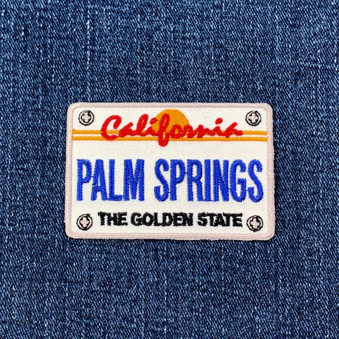 An embroidered patch of a California license plate, featuring the words "PALM SPRINGS" in blue letters and "California" in red script, all set on a white background on denim fabric.