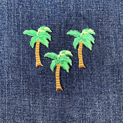 Three small embroidered palm trees with green fronds and tan trunks arranged on blue denim fabric.