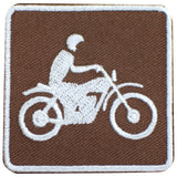 A square embroidered patch featuring a white silhouette of a person riding a motorcycle on a brown background, finished with a white stitched border.