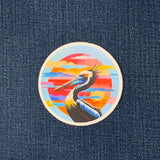 round sticker featuring a pelican with a colorful sky background displayed on a denim fabric