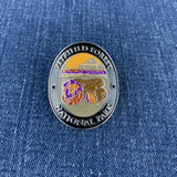 An oval enamel Walking Stick Medallion for Petrified Forest National Park, featuring a colorful landscape with a mountain and petrified wood, set against a blue denim background.